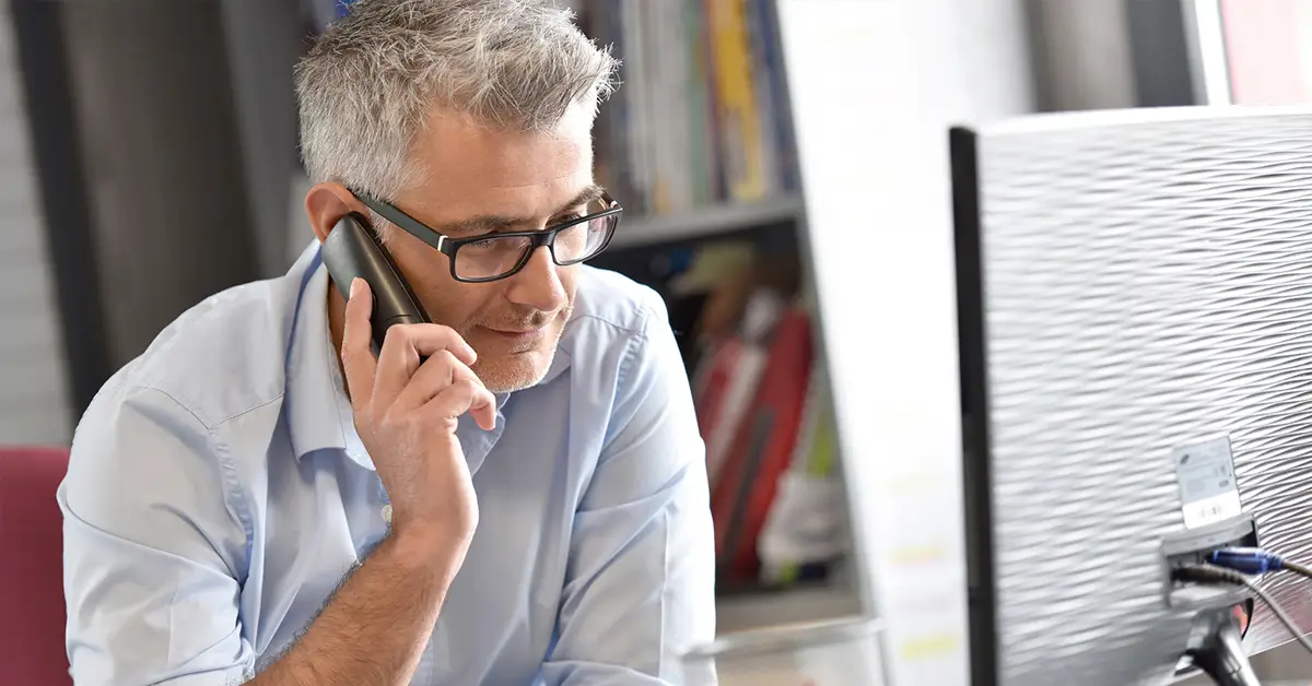 Man with glasses holding phone up to right ear while looking at computer monitor