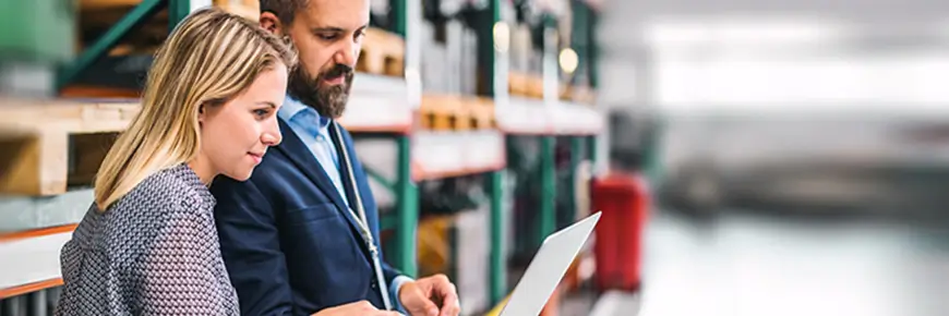 Man and woman standing up in a warehouse while looking at a laptop