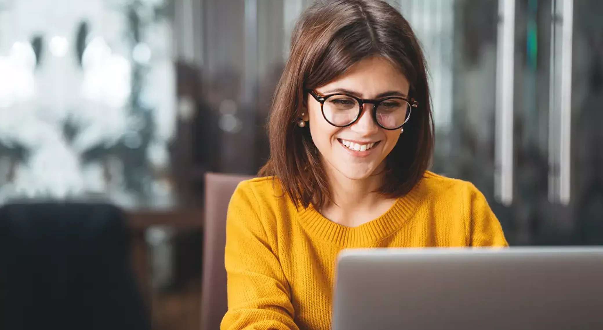 Woman with glasses smiling while looking down at laptop