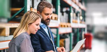 Man and woman standing up in a warehouse while looking at a laptop