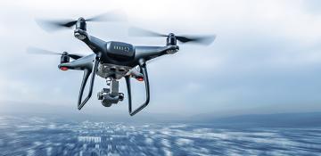 Image shows drone flying through the sky with a camera attached at the base. The drone is moving quickly and the background is blurred