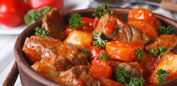 Image shows close up of meat stew. Made up of beef, carrot and potato in a tomato sauce with green herbs scattered on top.