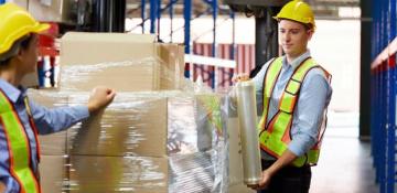 Image shows two people in a warehouse wrapping a pallet of cardboard boxes with plastic to secure for transportation. They are wearing high visibility safety equipment of a vest and a hard hat 