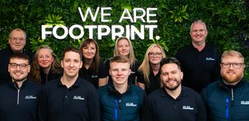 Image shows staff at company call We Are Footprint. It shows 11 men and women stood in front of a large wall of plants with the sign for the business lit up.